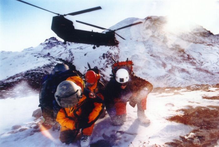 Chinook helicopter 90-00185 flies over Mount Rainier, Washington, while assigned to the Washington Army Reserve. Members of the National Park Service shield their faces from the sting of the ice and snow stirred up by rotor blades of the helicopter that had just deposited them there. TheNational Park Service Rangers prepare to practice for high-altitude rescues. They're at the 9,000-foot level on the northeast side of Mount Rainier, at Camp Schurman.